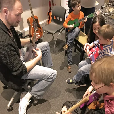An adult tutor and children playing ukuleles and guitars in a group music lesson