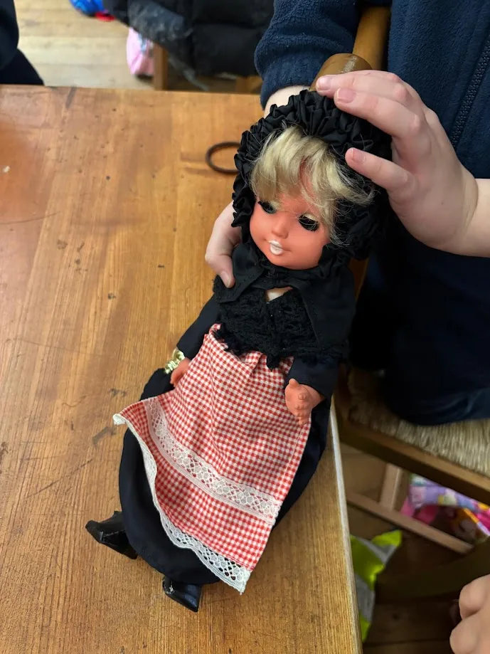 A hand holds a doll in a dark dress with a red and white checked apron upright on a wooden table.