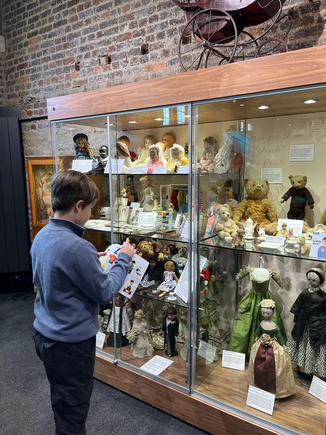 A boy writes on a worksheet in front of a tall glass display case of dolls and teddy bears in the Toy Museum.