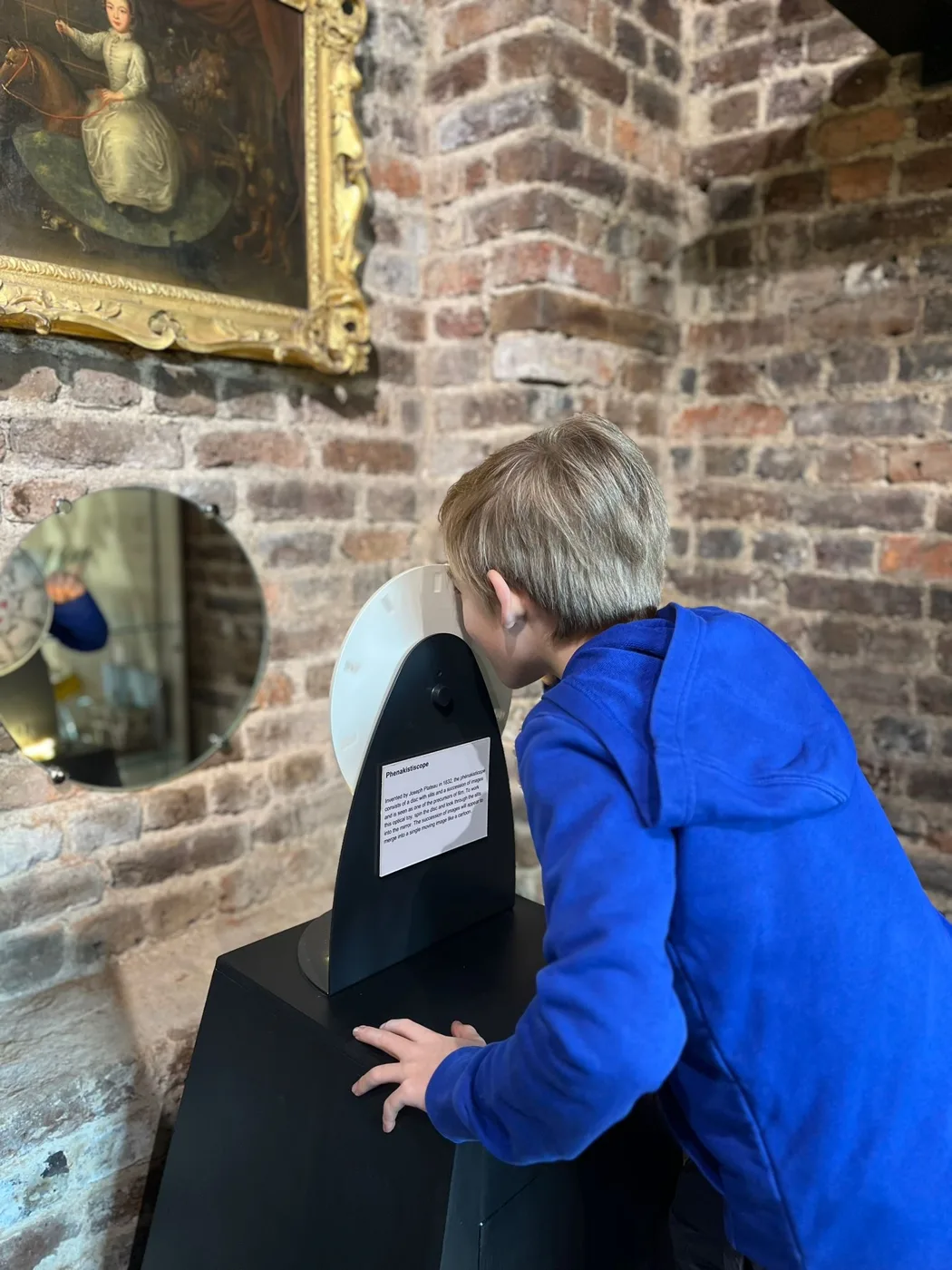 A boy in a blue fleece leans in to look closely at an exhibit beside a brick wall.