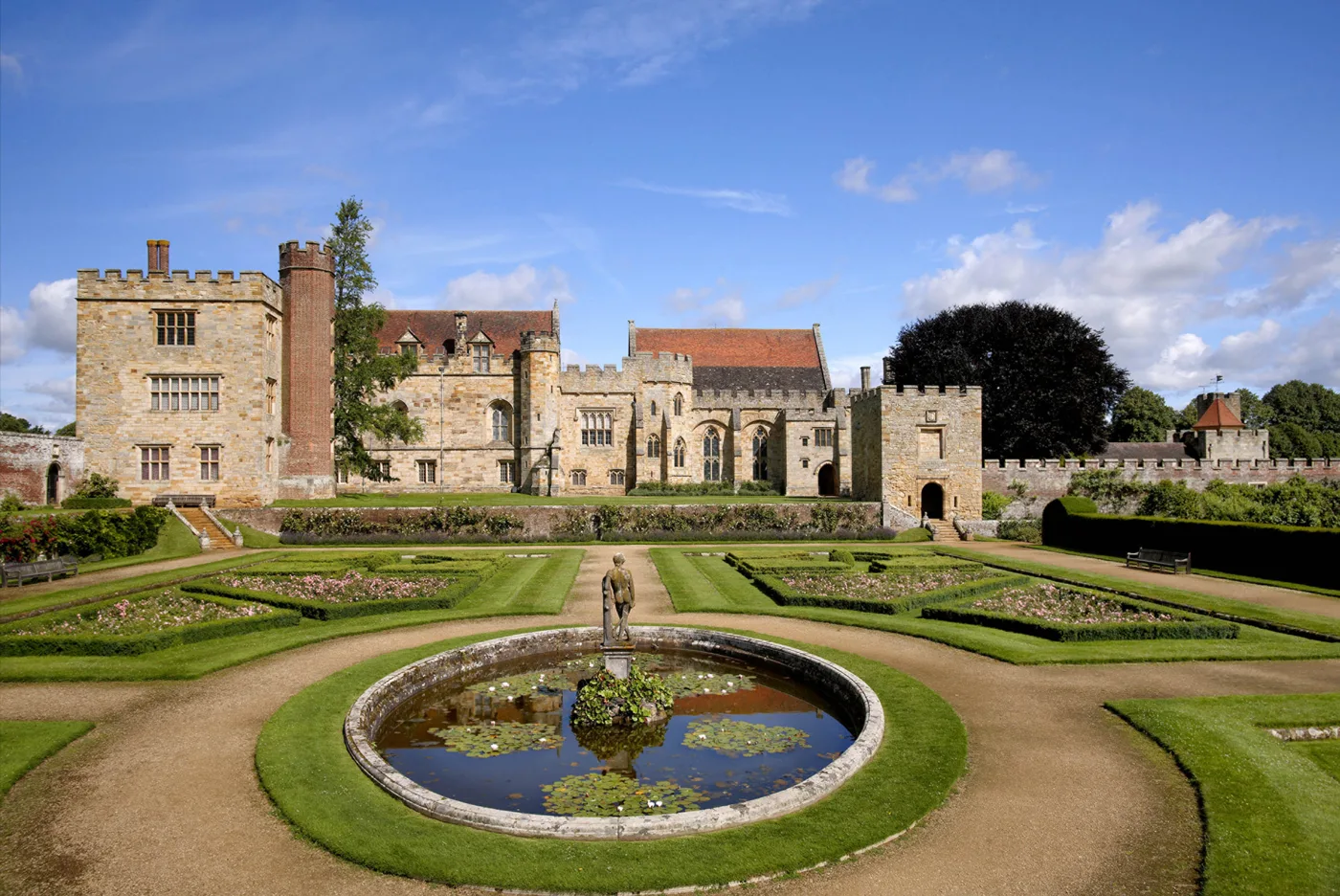 The south front of Penshurst Place, a long stone house with towers and battlements, seen across a formal garden with a round pond.