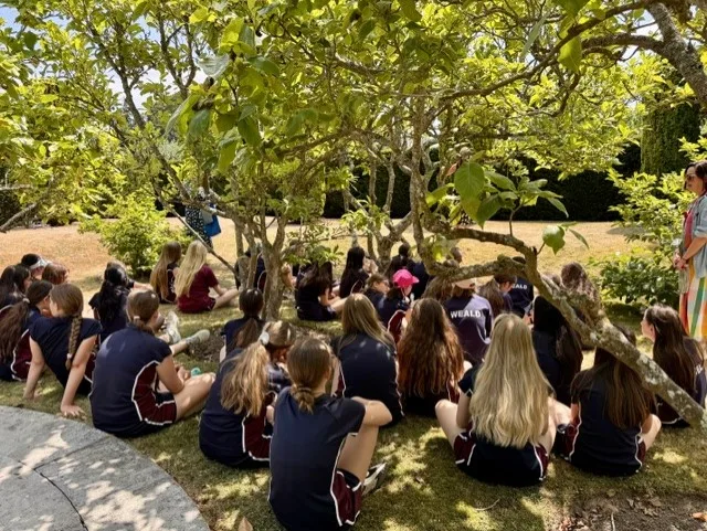 A large group of pupils in school uniform sitting on the grass in the shade of a tree in the gardens at Penshurst Place.