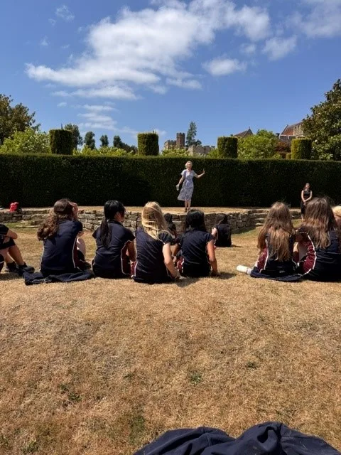 Pupils in school uniform sit on the lawn beside a tall clipped hedge, facing a person standing in front of them.