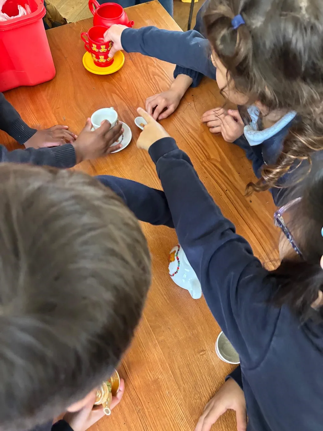Young pupils around a wooden table reaching for a toy tea set during a workshop.