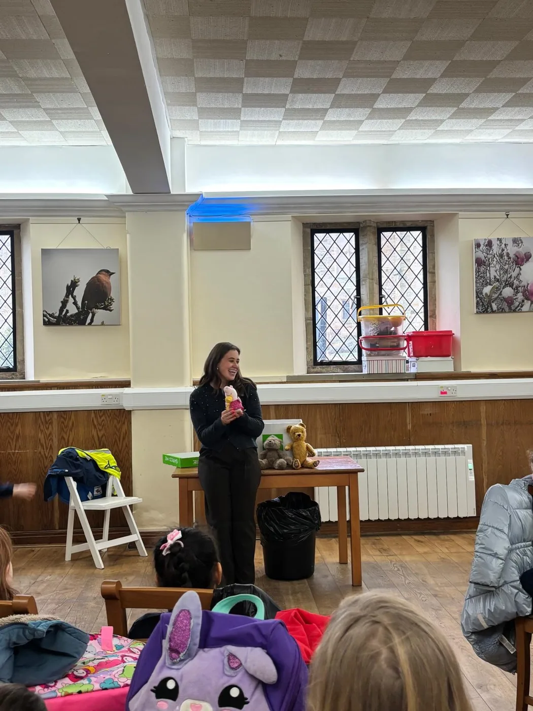 A session leader stands at the front of a hall with leaded windows, holding up a toy, as pupils with backpacks and high-visibility vests sit and watch.