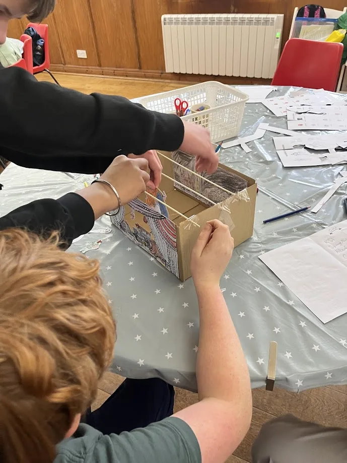 Pupils' hands at a table building a small cardboard theatre, with craft materials around them.