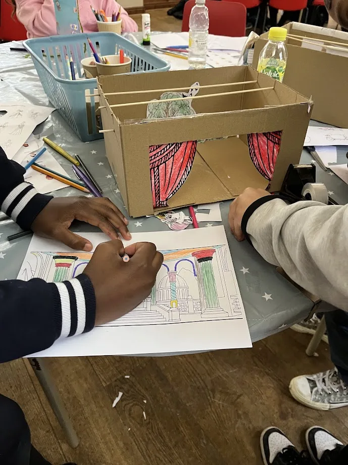 A cardboard toy theatre with paper curtains on a table, as pupils colour in designs beside it.