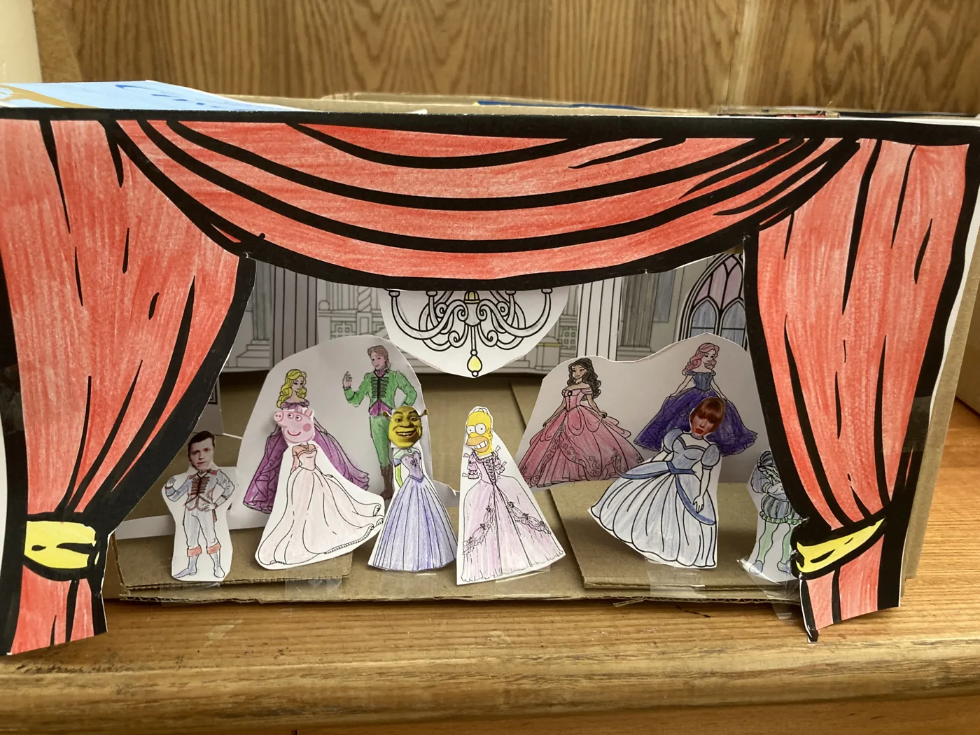 A finished cardboard toy theatre with red paper curtains and hand-drawn paper figures in ballgowns on its stage.