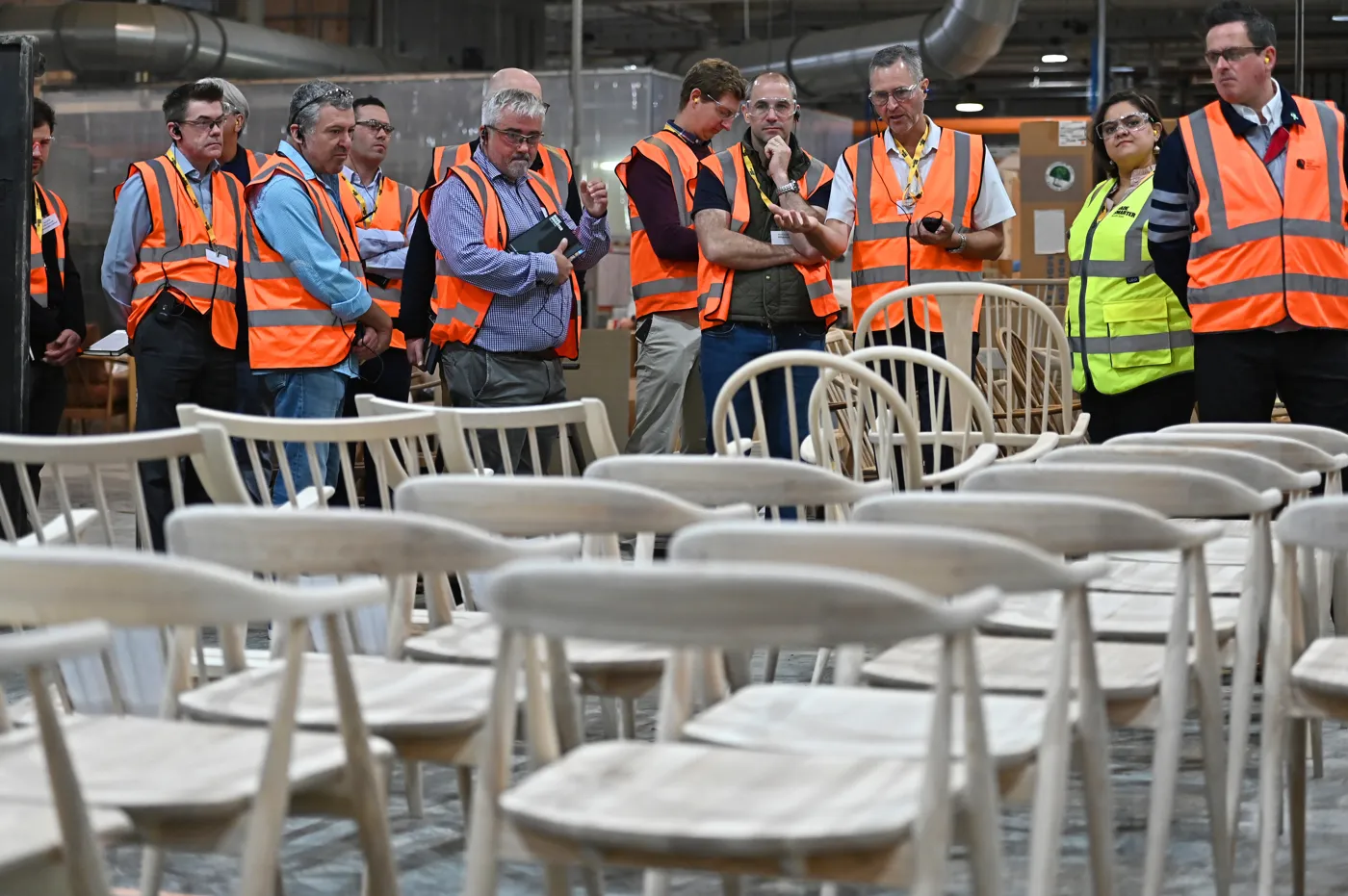 Delegates in orange high-visibility vests stand behind rows of unfinished pale wooden chairs on the factory floor at Ercol.