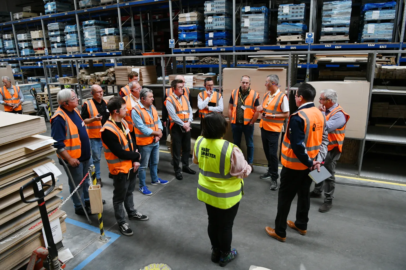 A group in high-visibility vests gathers in a warehouse aisle lined with racks of timber and boards, listening to a guide.