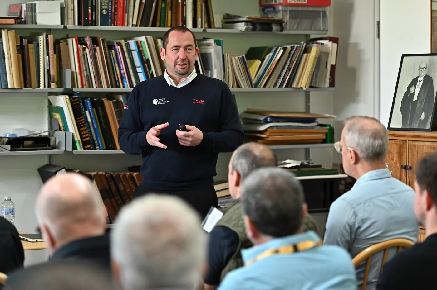 Steve Nelson, in a navy Toyota Lean Management Centre jumper, speaks to seated delegates in front of shelves of books.