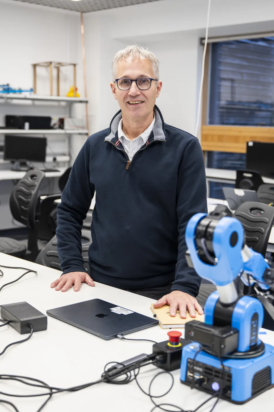 Ian Peers stands at a desk next to a small blue robotic arm, a laptop and cables.